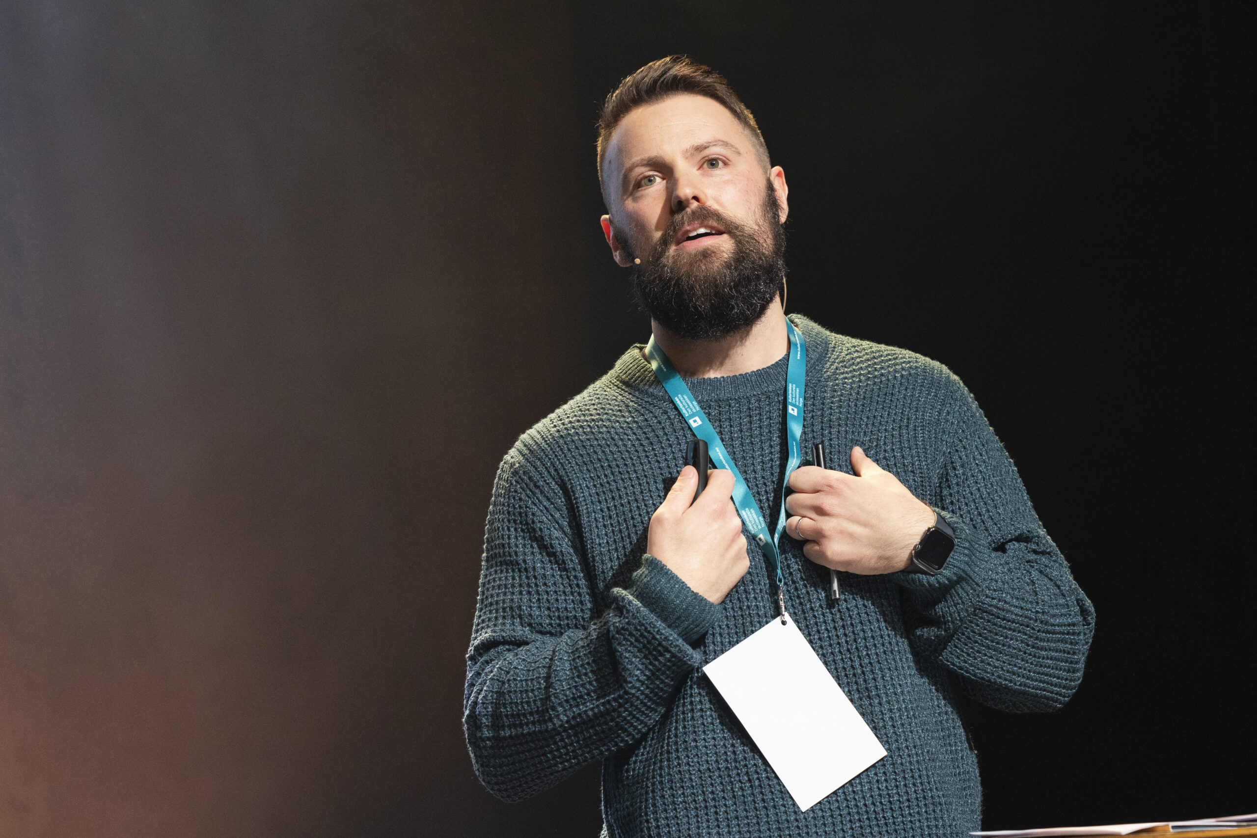 A white man with brown hair and beard standing with his hands up to his chest. He is wearing a green jumper and has a lanyard on his neck.