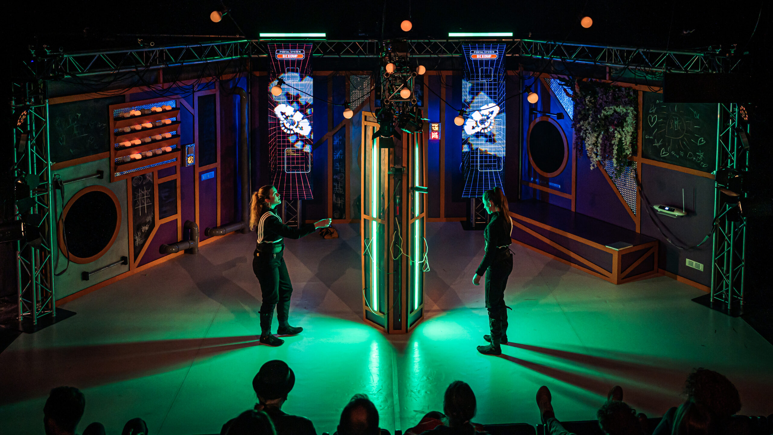 Two performers in dark jumpsuit costumes on a sci-fi style set with lots of neon coloured lighting and two big screens with computer graphics.