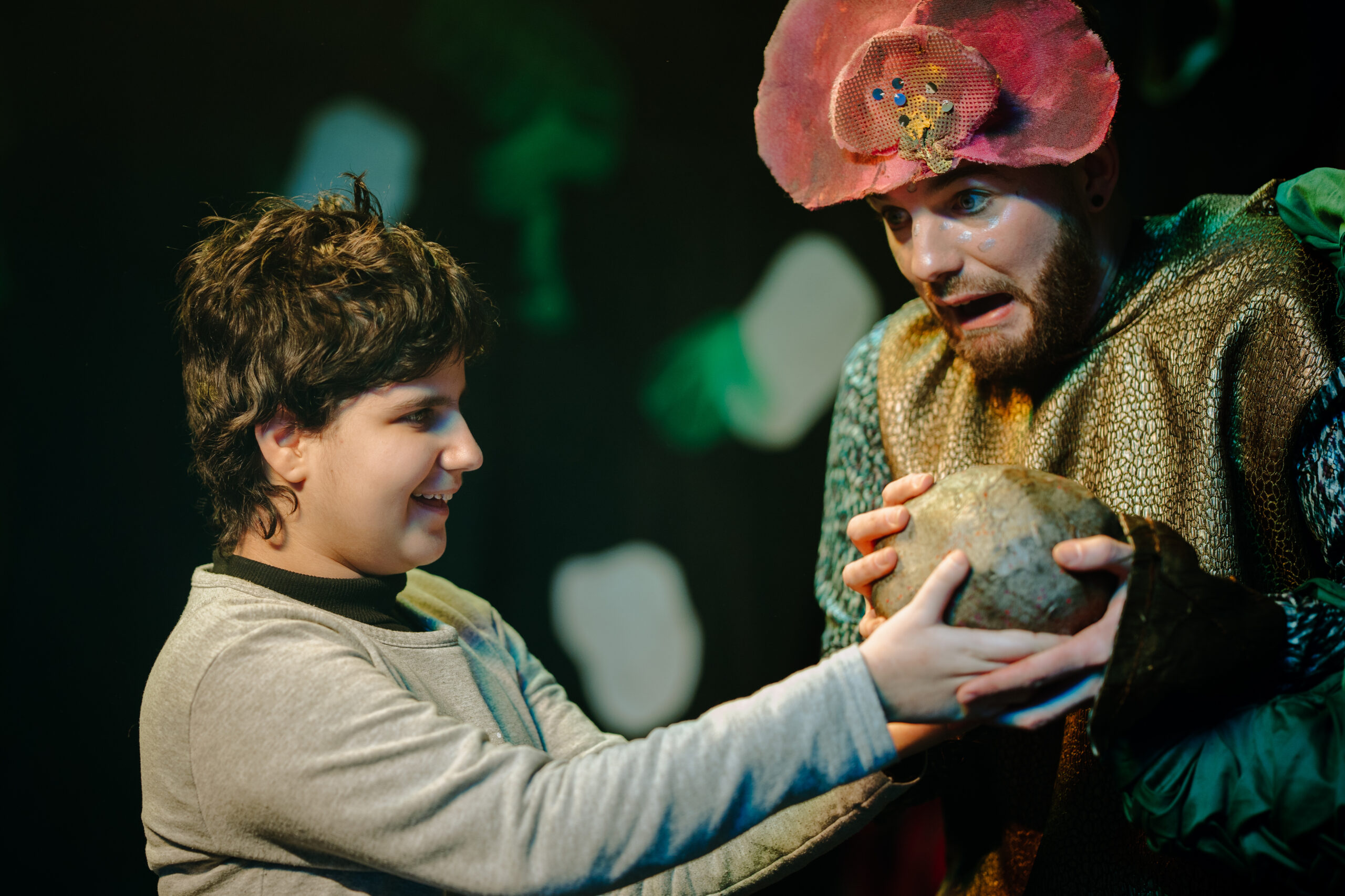 A young audience member smiles as they and a performer wearing a headpiece and costume inspired by leaves hold a grey prop stone together.