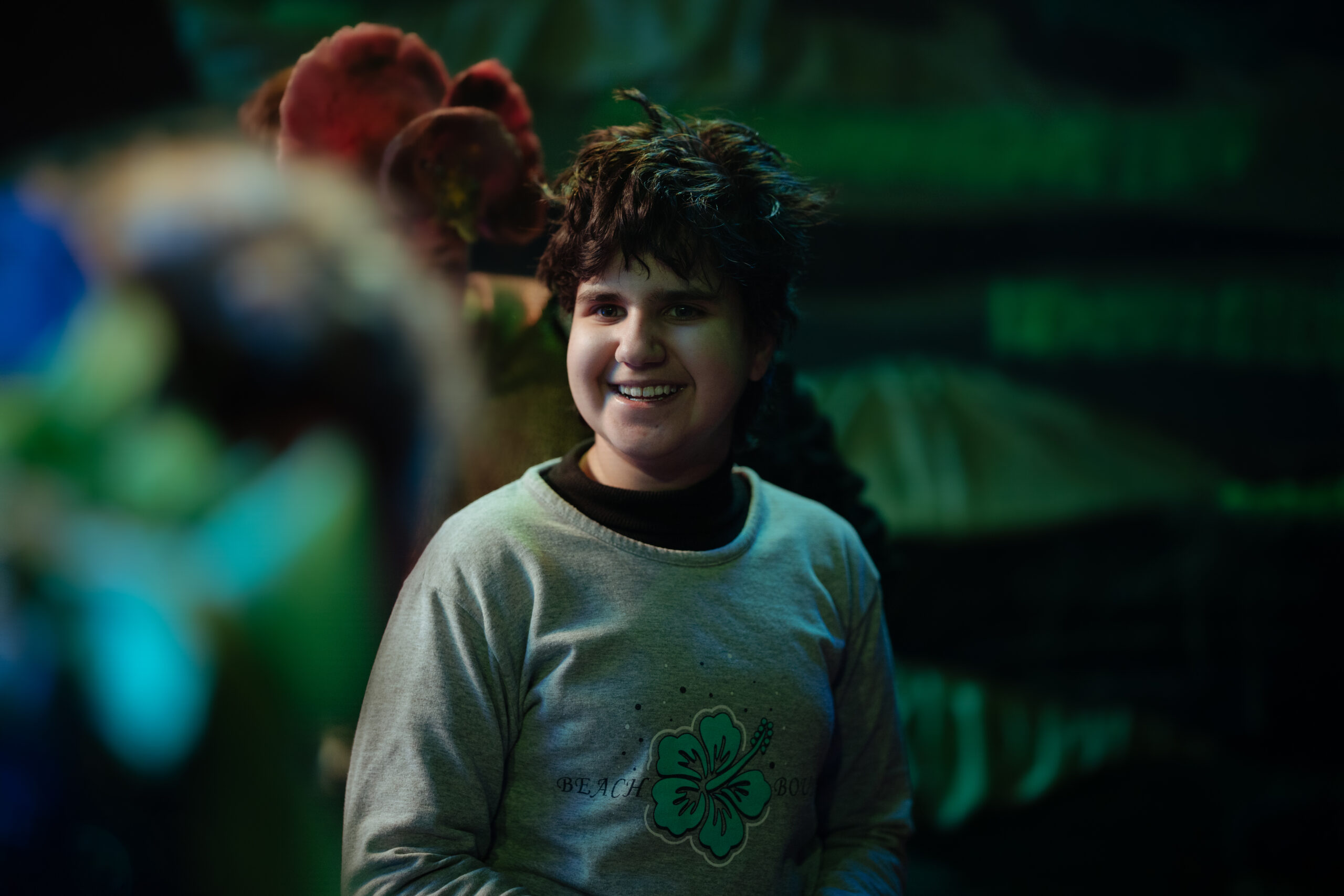 A young person smiles at a performer off-camera. They are in front of a textured green backdrop and there is a performer in a red costume moving behind them.