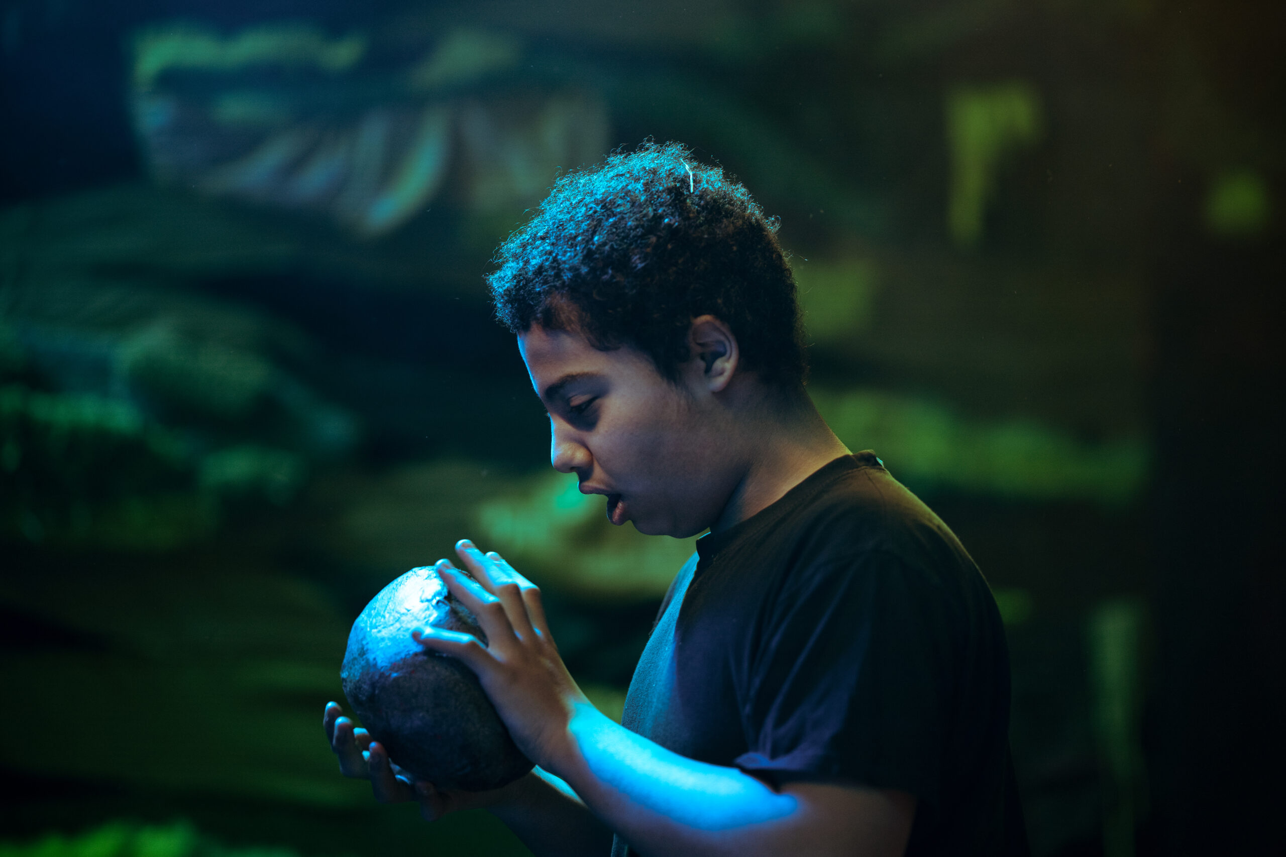 A young audience member holds a theatre prop in the shape of a large stone with a textured grey surface. Behind them is a textured green background and they are under blue lighting.
