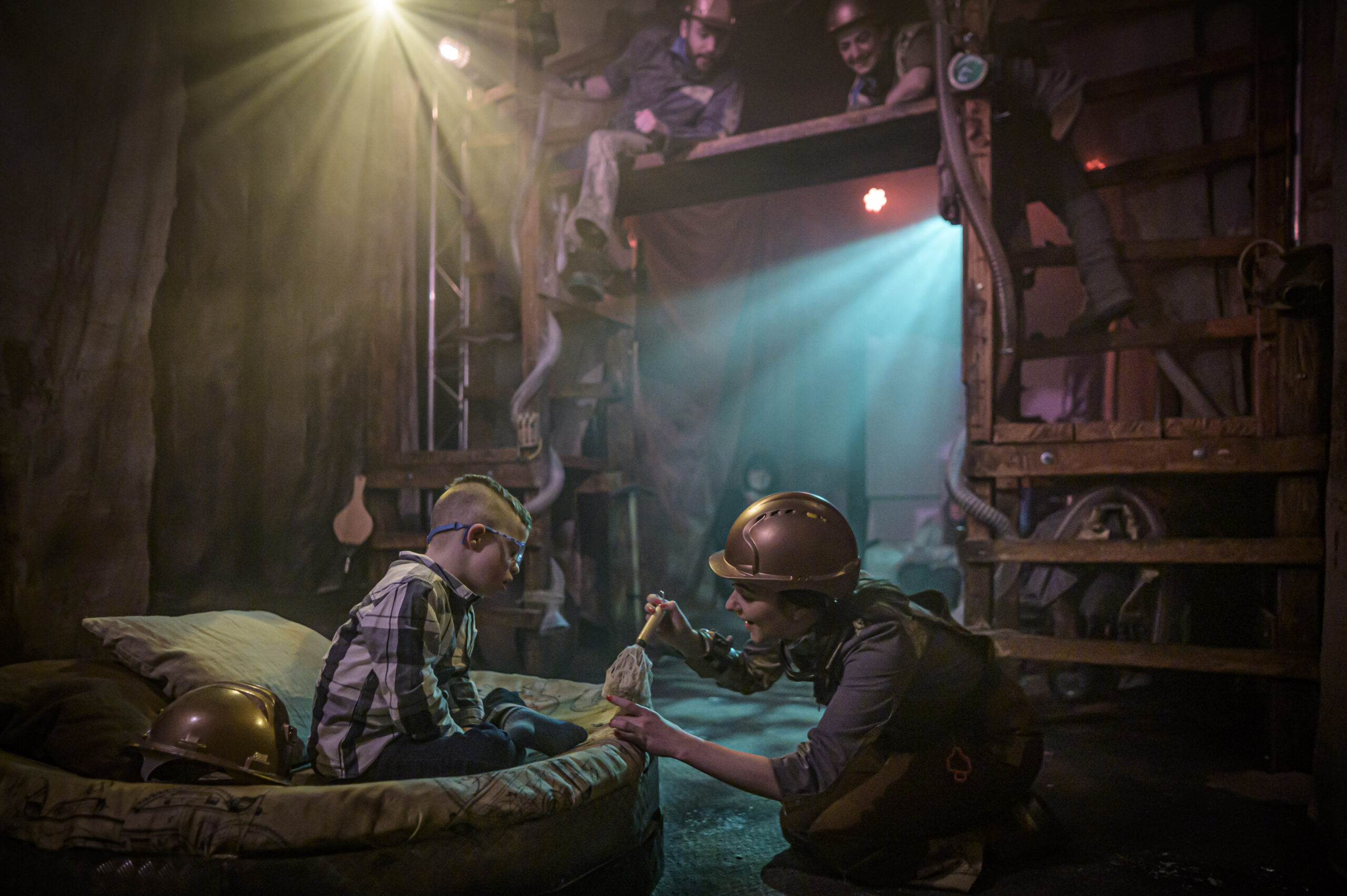 Three performers and a young audience member on the set of Hollow, which is a dark space inspired by an underground mine. One of the performers is holding a small brush, and the audience member is sitting beside them, watching the action.